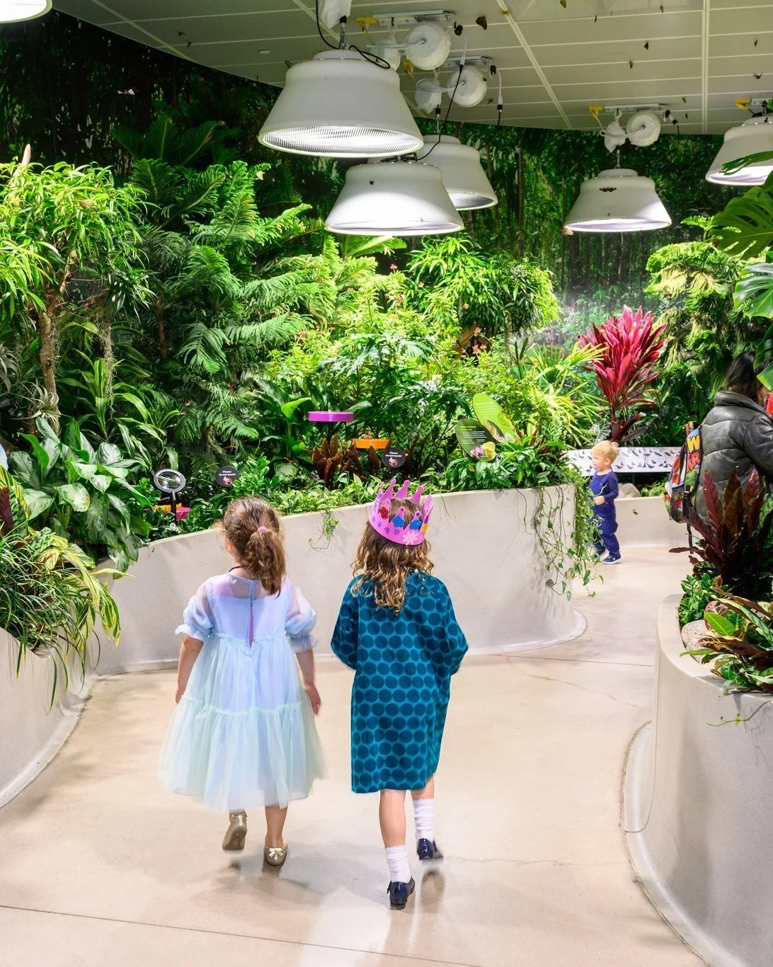 A photo of two children walking through the Butterfly Vivarium. One of them is wearing a pink paper crown.