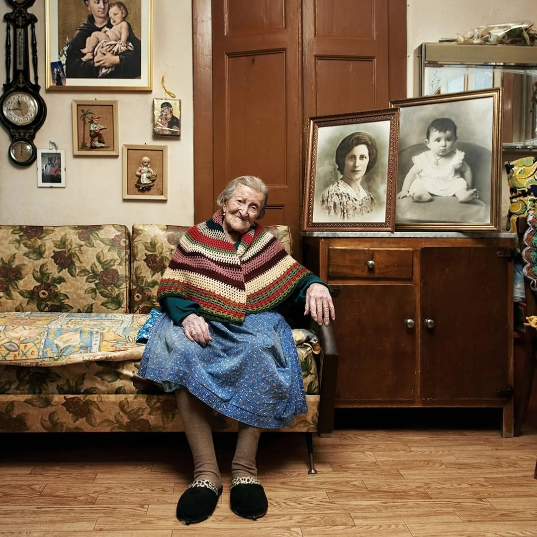 A photo of Emma Morano sitting on a floral print sofa. She is wearing a shawl and a blue dress. She is surrounded by an eclectic mix of objects, including two framed portrait photos.